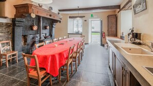 Ein Essbereich mit einem langen Tisch, der mit einer roten Tischdecke bedeckt ist, Holzstühle, ein Kamin aus Stein und eine Küchenzeile inklusive Spüle und Herd.