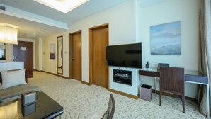 Una camera d'albergo con televisione a schermo piatto, una scrivania con un computer, un divano e un tavolino. Si vedono tre porte e un quadro appeso al muro.