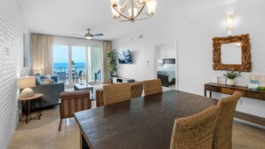 A spacious dining area with a large wooden table and wicker chairs, a chandelier, and a view of the ocean through large windows.