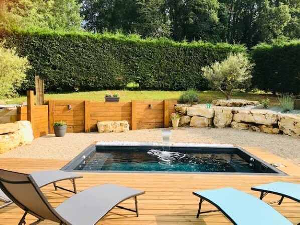 A hot tub surrounded by wooden deck chairs, a stone wall, and a hedge.