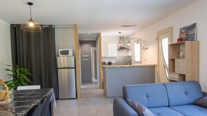 A modern living room with a grey sofa, a dining table with chairs, a kitchen area with a fridge, microwave, and cabinets, and a hallway leading to another room.
