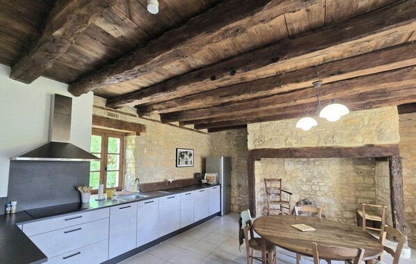 A rustic kitchen with a stone wall, wooden ceiling beams, a large island, and a dining table with chairs.