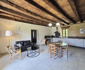 A spacious living area with a black leather sofa, a round coffee table, and a dining table with chairs. There is a kitchen with a stone wall and a window.
