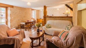 A cozy living room with a sofa, two armchairs, a wooden coffee table, and a dining table with chairs. There is a kitchen area with a wooden counter and a window.