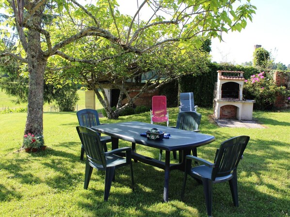 Un patio al aire libre con una mesa y cuatro sillas, rodeado de abundante follaje y un horno de ladrillo.