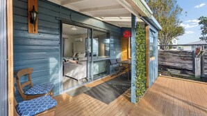 Un espacio exterior cubierto con una terraza de madera, una zona de estar con una silla y mesa, y un ventanal corredizo que da a un espacio interior.