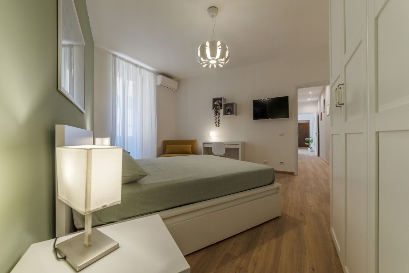A bedroom with a bed, bedside table, lamp, and a TV on the wall.