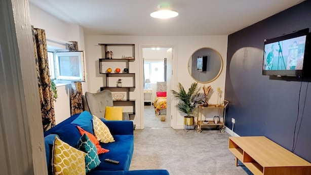 A living room with a blue sofa, a round mirror, a TV mounted on the wall, and a small wooden table. - Mother of Stockton (Stockton-on-Tees)