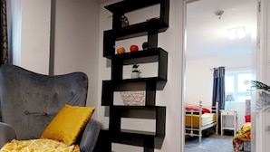A room with a grey upholstered armchair, a yellow throw blanket, and a black shelving unit with decorative items. - Mother of Stockton (Stockton-on-Tees)