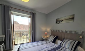 Ein Schlafzimmer mit Bett, Nachttisch, Stuhl, einem Balkon mit Blick auf ein Gebäude und einem Wandbild.