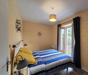 A bedroom with a bed, a bedside table, a lamp, a wall-mounted shelf, a window with curtains, and a view of greenery outside.