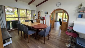 Un coin repas avec une table et des chaises en bois, une vue sur l’extérieur grâce à de grandes fenêtres, une horloge au mur et une porte donnant sur une autre pièce.