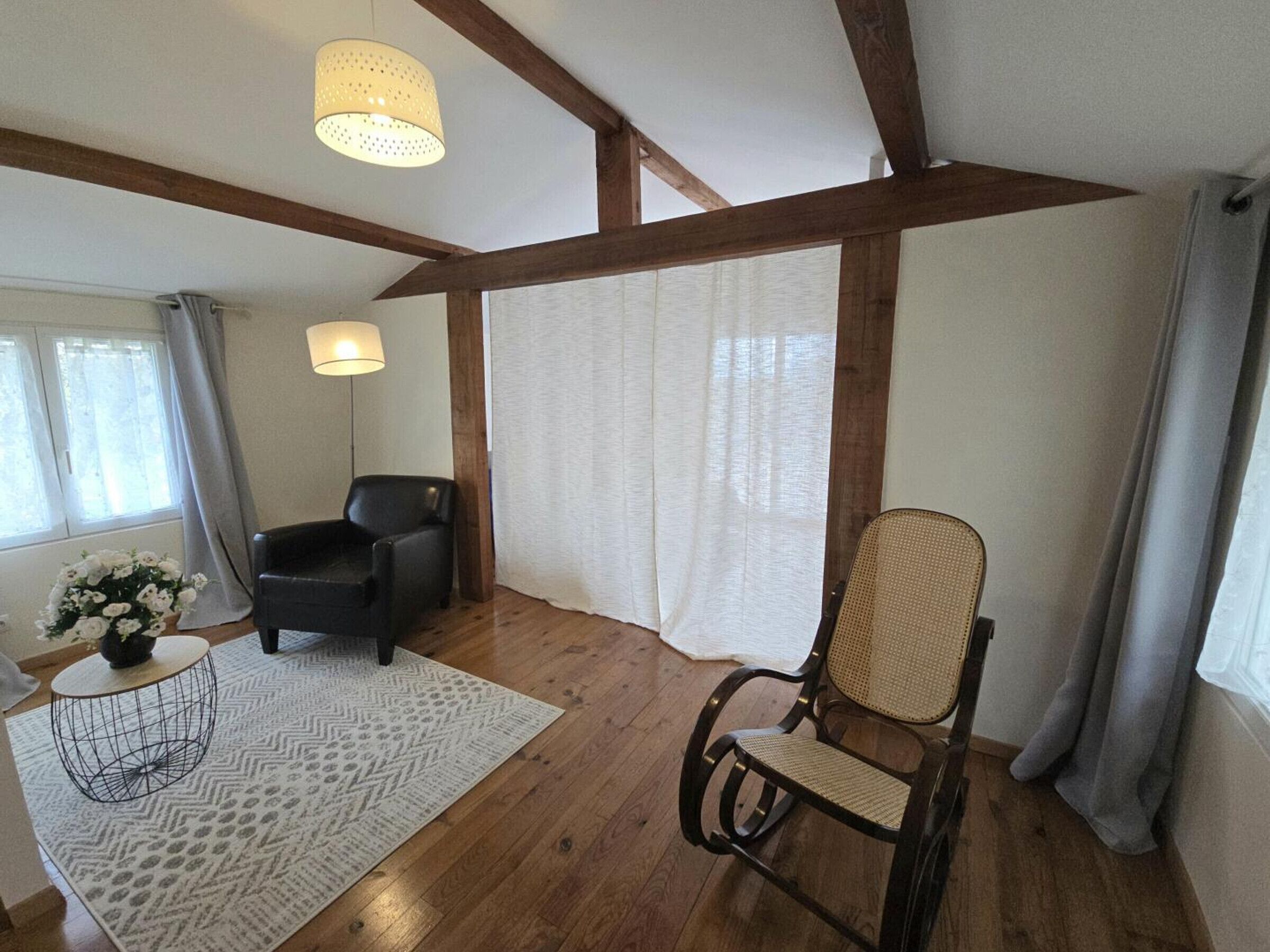 A room with wooden beams, a black leather chair, a woven chair, a small round table with a lamp, and a patterned rug.