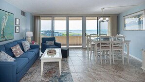 A living room with a blue sofa, a coffee table, and a dining area with a table and chairs. - Beautiful sunset and ocean view from 2 Bedroom Condo with Free WiFi and an Outdoor Pool Located in Midtown Only Steps to Beach! Vacation at the Shore with Shoreline Properties (Ocean City)