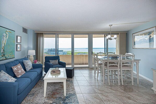 A living room with a blue sofa, a coffee table, and a dining area with a table and chairs. - Beautiful sunset and ocean view from 2 Bedroom Condo with Free WiFi and an Outdoor Pool Located in Midtown Only Steps to Beach! Vacation at the Shore with Shoreline Properties (Ocean City)