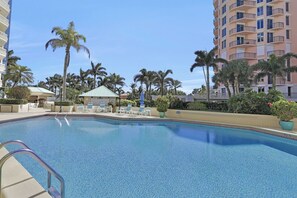 A swimming pool with lounge chairs and umbrellas, surrounded by palm trees and a multi-story building.