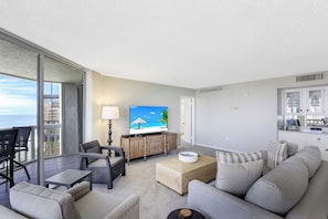 A modern living room with a large TV, comfortable seating, a dining area, and a view of the ocean. - Beachfront Condo Building | Pool, Tennis & Views (Marco Island)