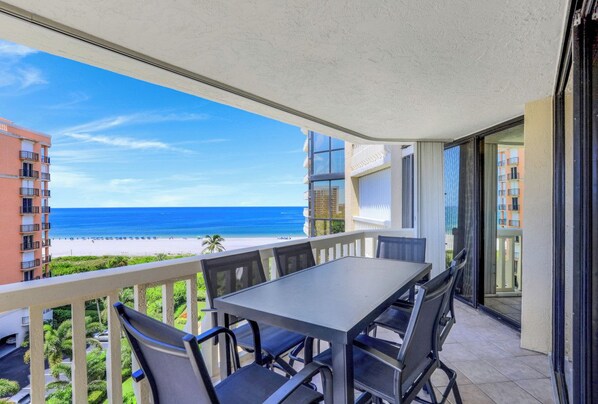 A balcony with a table and chairs, overlooking a beach and ocean.