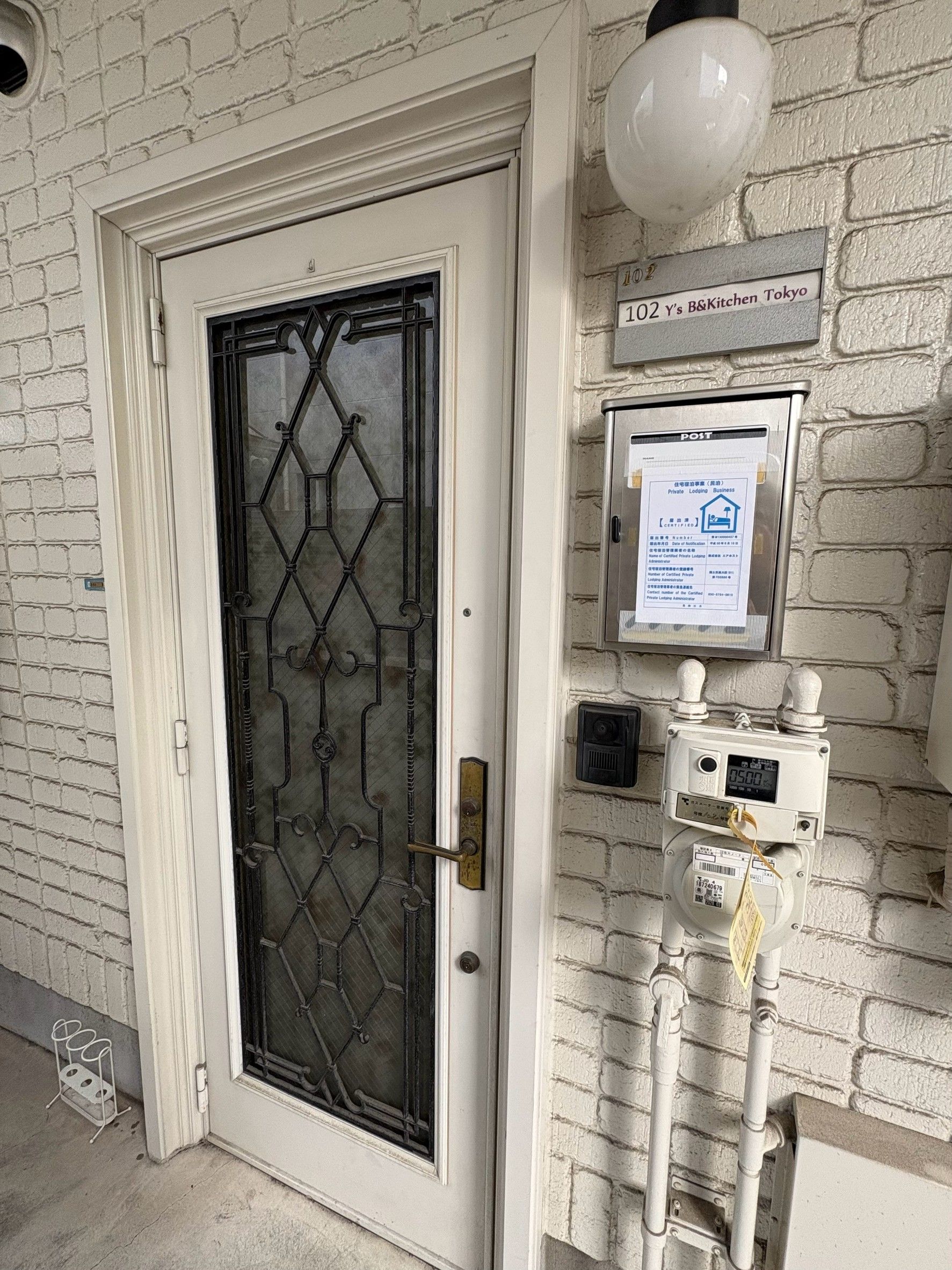 Une porte ornée d’une grille métallique décorative, une enseigne fixée au mur indiquant qu’il s’agit d’un établissement proposant des chambres en location, et un compteur d’eau ou d’électricité.