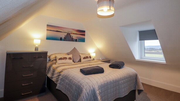 A bedroom with a bed, a dresser, a painting, a window, and a ceiling light fixture. - Nanville (Beauly)