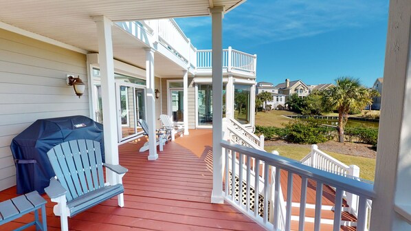 A covered porch with Adirondack chairs, a grill, and a view of a residential area with houses and a pond.