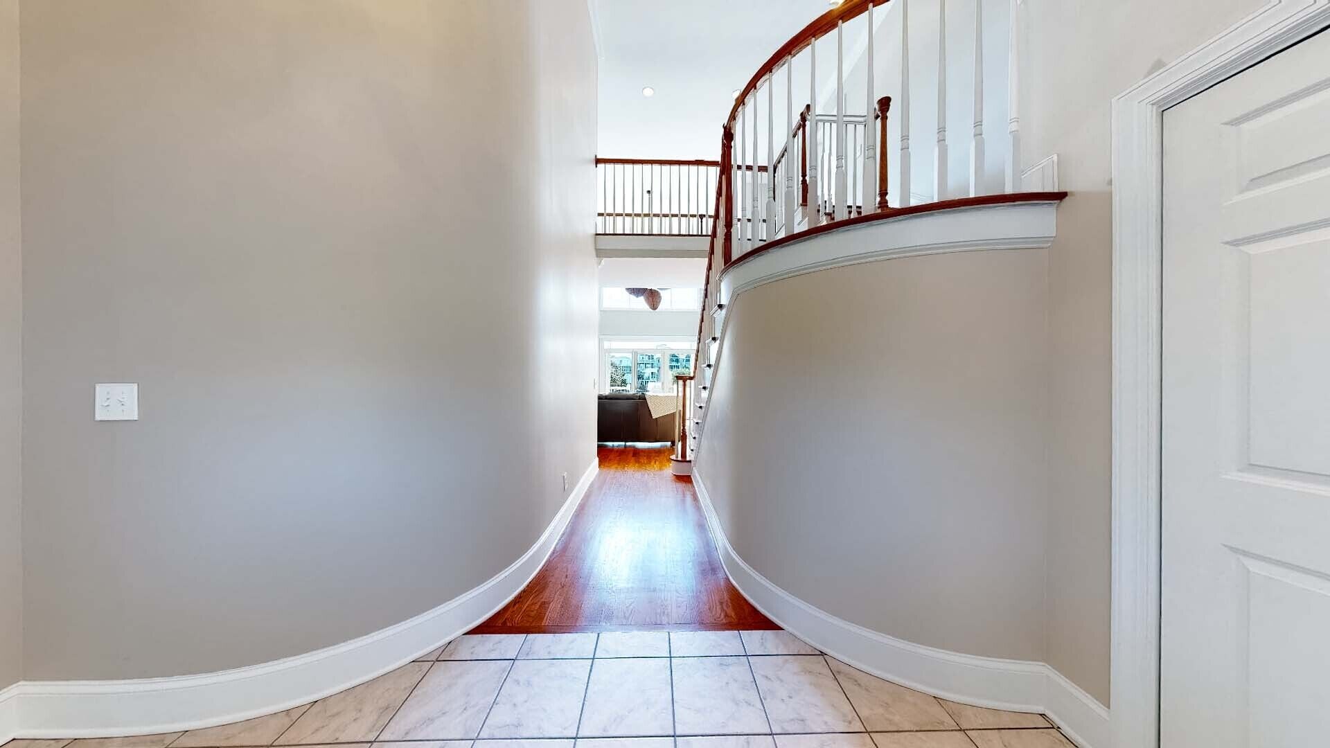 A hallway with a curved floor, white walls, and a wooden staircase with a railing.