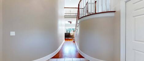 A hallway with a curved floor, white walls, and a wooden staircase with a railing.