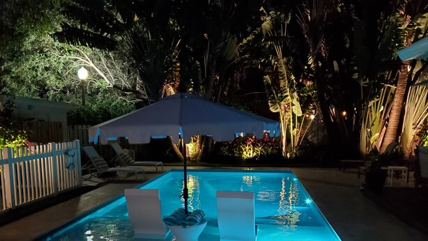 Una piscina rectangular con agua cristalina, rodeada por una terraza de madera y una valla blanca. Hay sillones de descanso y una sombrilla azul que proporciona sombra.