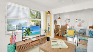 Una sala de estar moderna con televisor mostrando una escena de playa, un comedor con mobiliario de mimbre y un espejo grande.