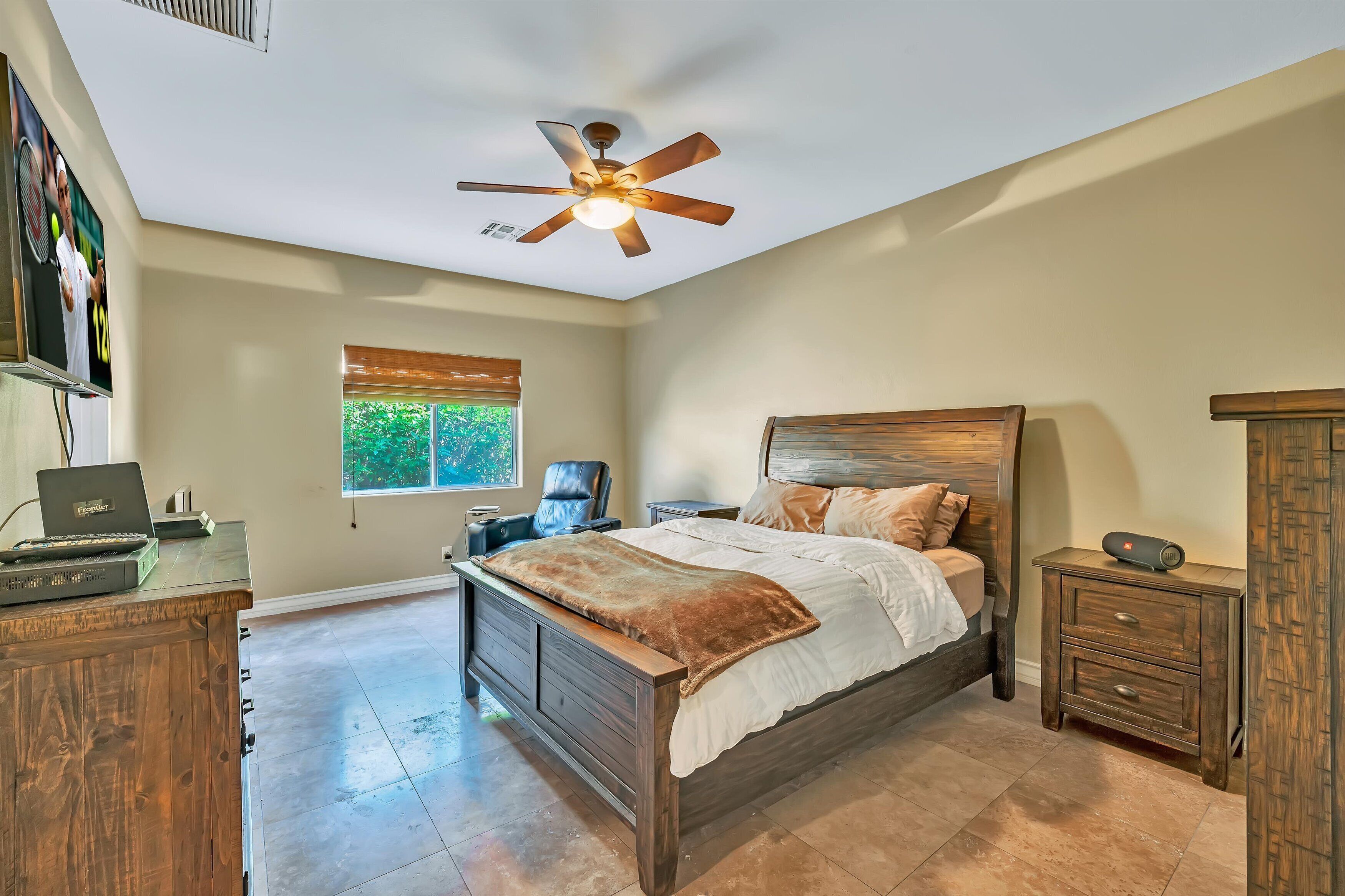 A bedroom with a bed, a desk with a laptop, a ceiling fan, a window, and a nightstand.