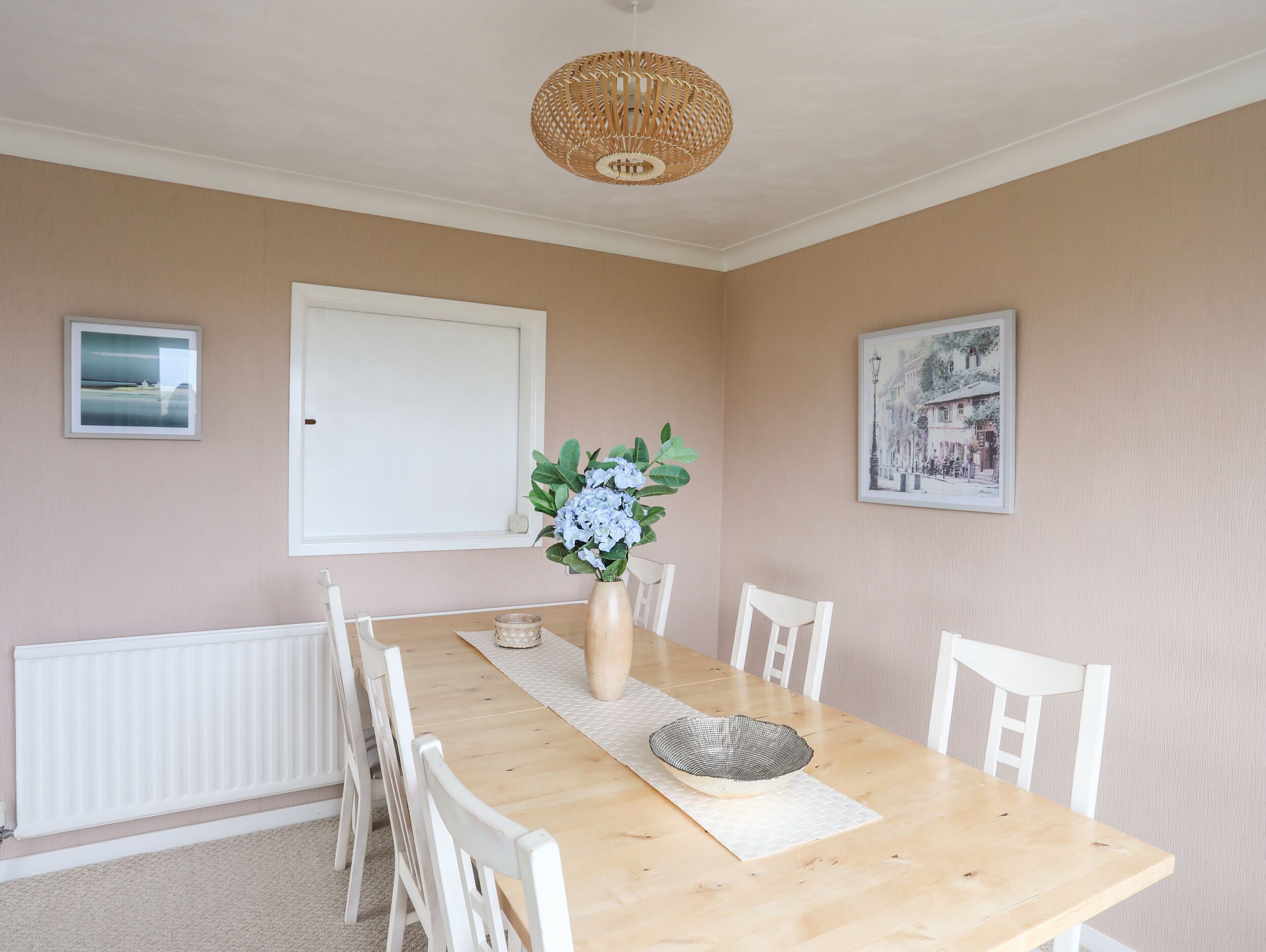 A dining room with a wooden table, white chairs, a vase with flowers, and two framed pictures on the walls.