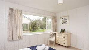 A bedroom with a large window, a bed with a blue bedspread, a white chair, a dresser, and a framed picture on the wall. - Ty Bedw (Tyn-y-Gongl)