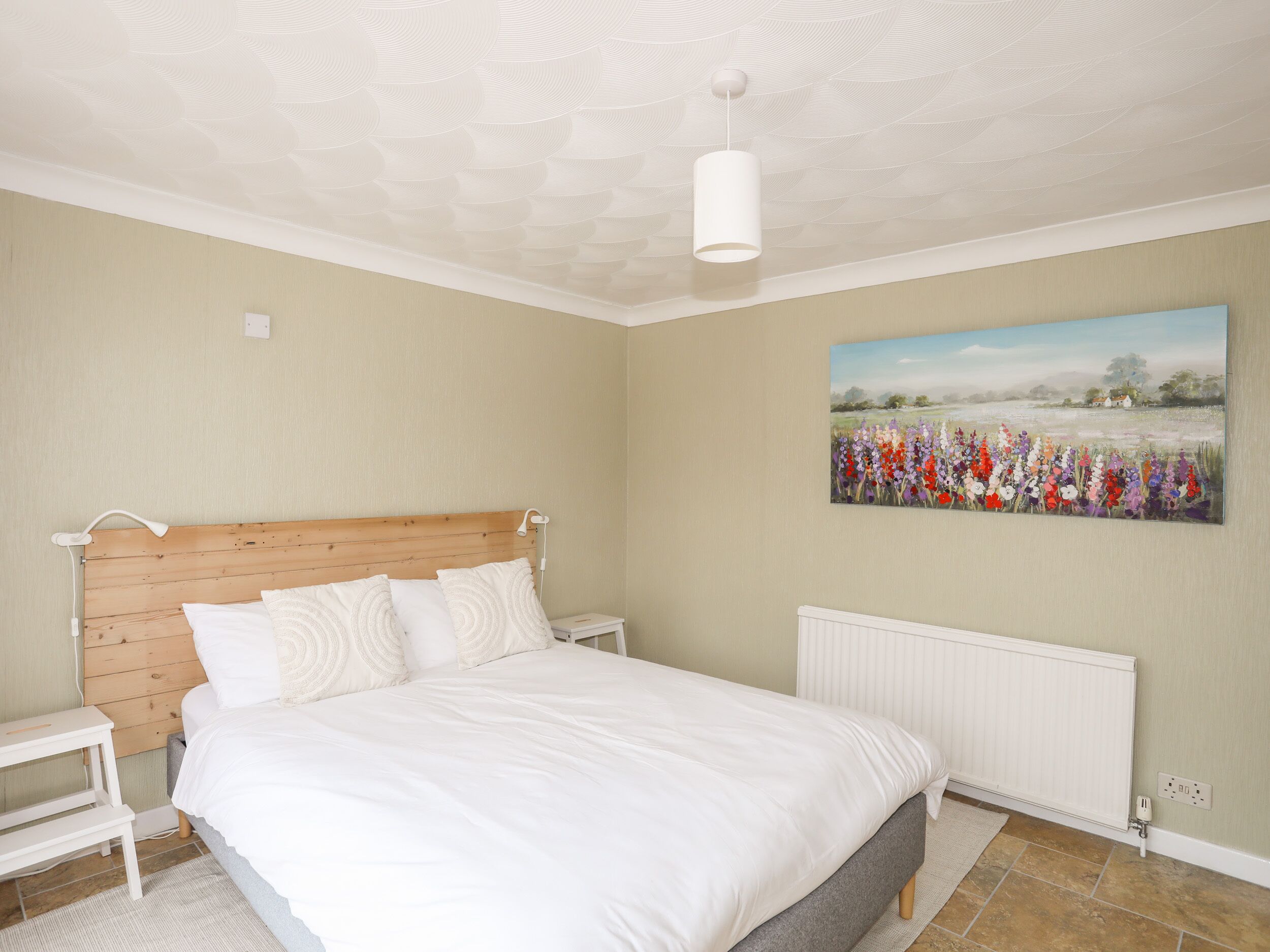 A bedroom with a wooden headboard, white bedding, a nightstand, a bedside table, a radiator, and a painting of flowers on the wall.