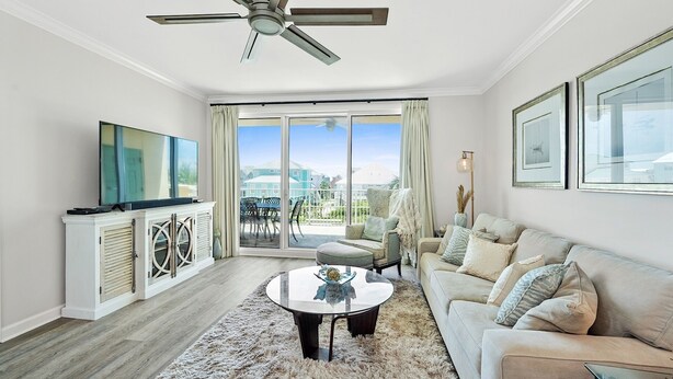 Ein geräumiges Wohnzimmer mit Deckenventilator, Fernseher, Essbereich mit Tisch und Stühlen, einer Couch und großen Fenstern mit Blick nach draußen.