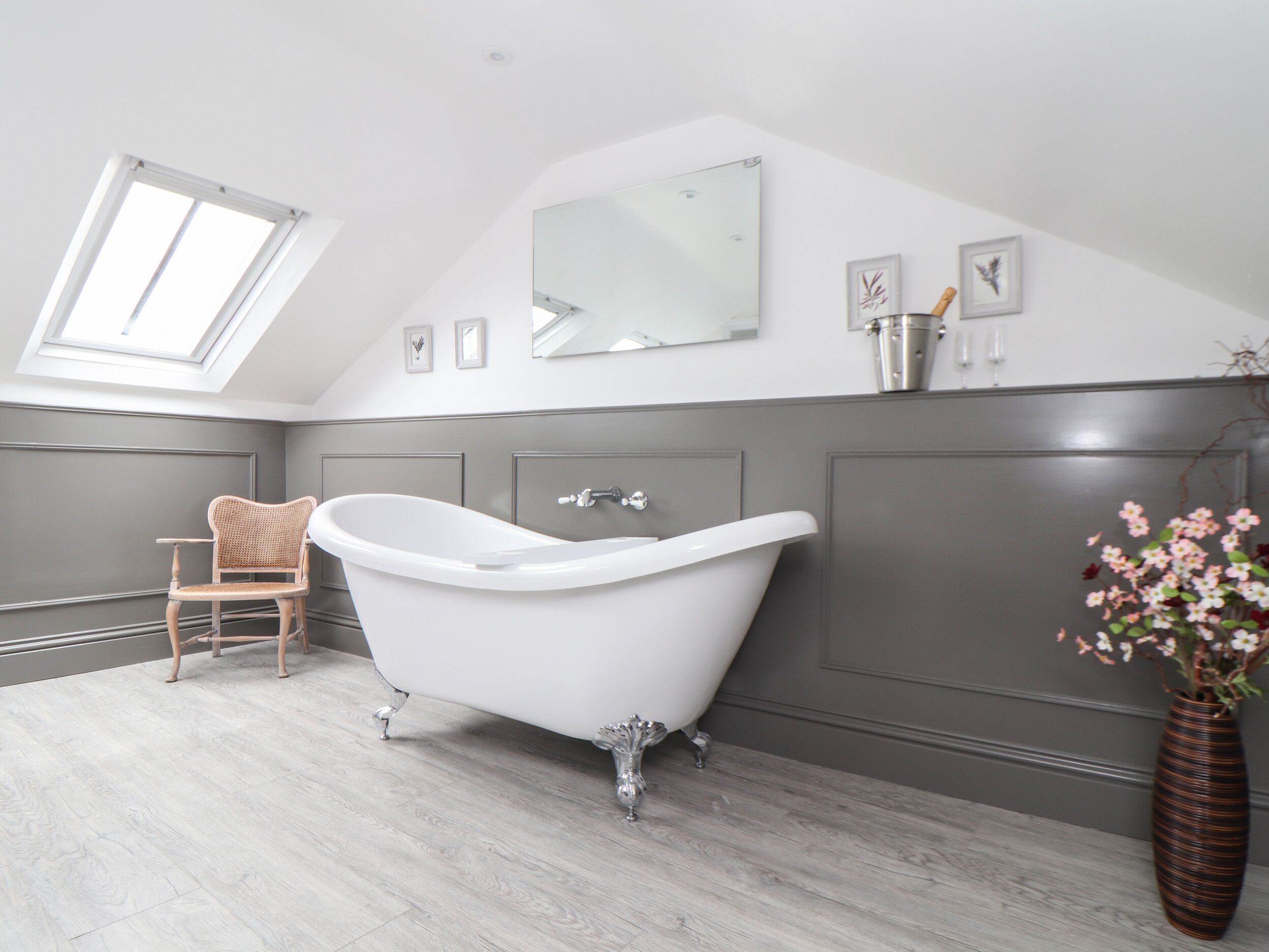 Une salle de bain moderne avec une baignoire sur pieds, une chaise en bois, un grand miroir et une lucarne.