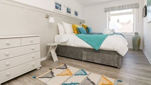 A bedroom with a bed, dresser, bedside table, lamp, window, and artwork on the wall. - Rock Pool House (Helston)