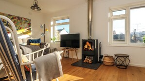 A cozy living room with a fireplace, wooden flooring, a TV, and a view of the city through large windows. - Rock Pool House (Helston)