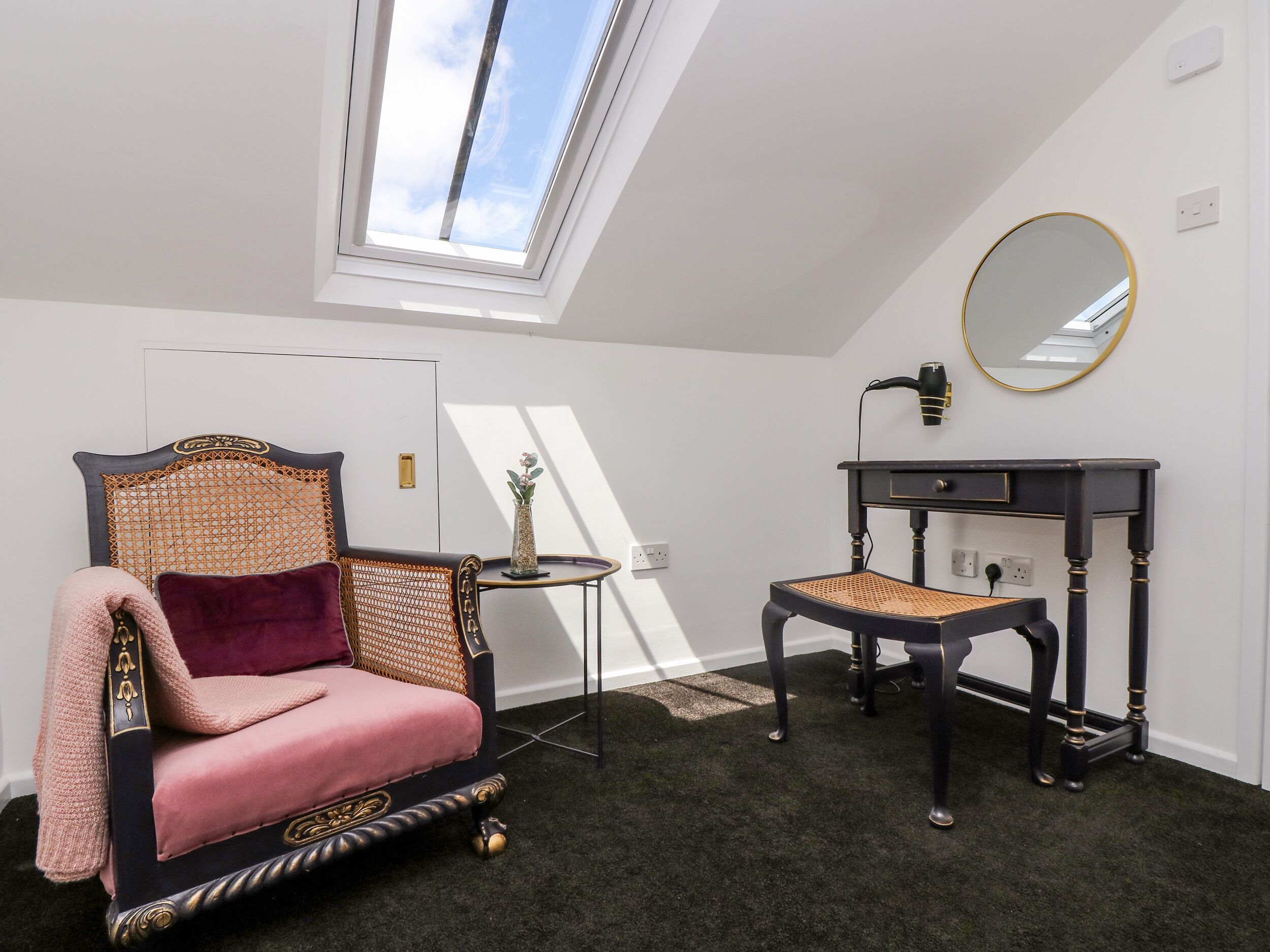 A room with a high ceiling, a skylight, a chair with a cushion, a small table with a vase, a desk with a lamp, and a round mirror on the wall.