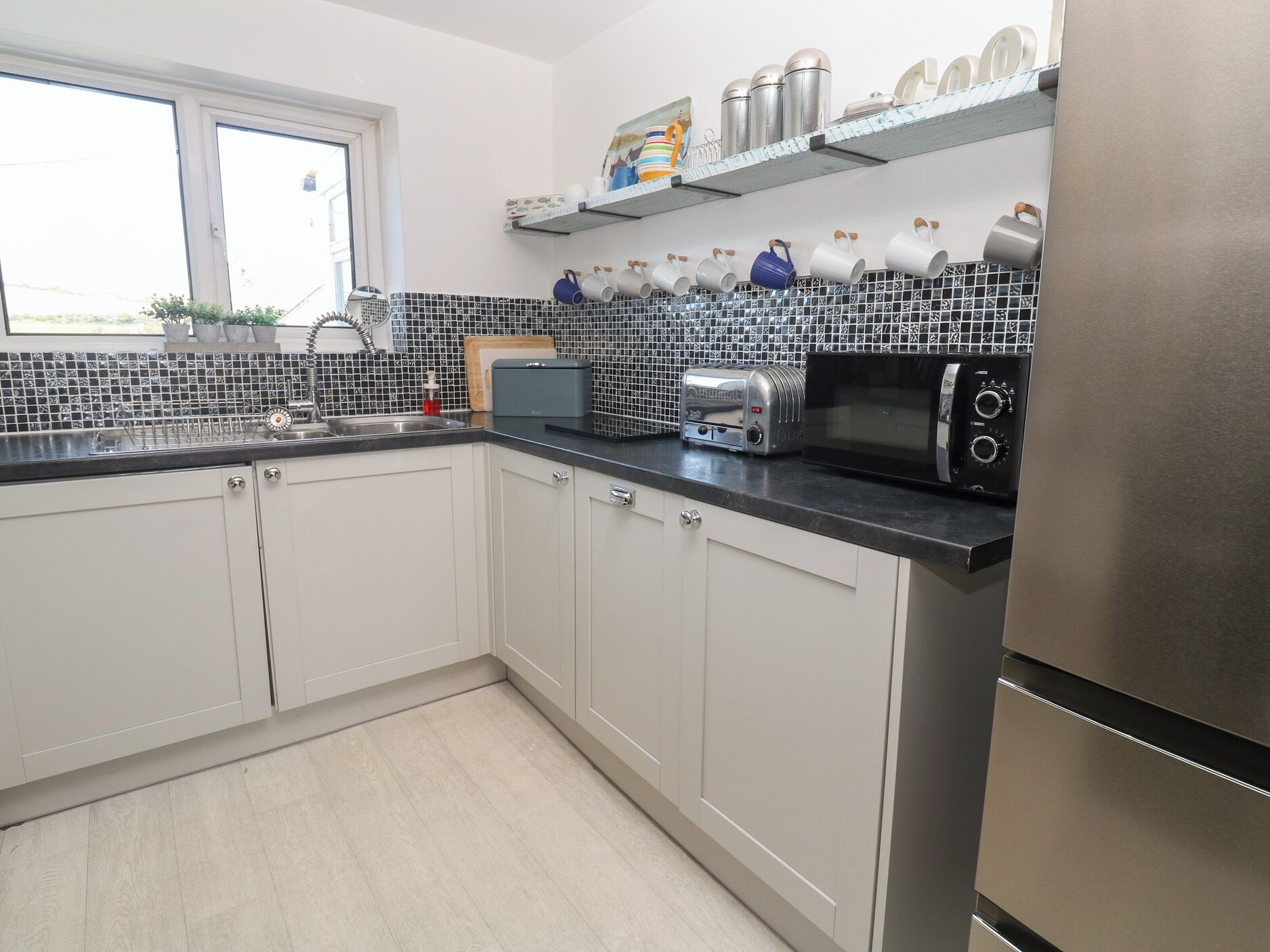 A kitchen with white cabinets, a black countertop, a stainless steel refrigerator, a microwave, a toaster, and a window with a view of the outdoors.