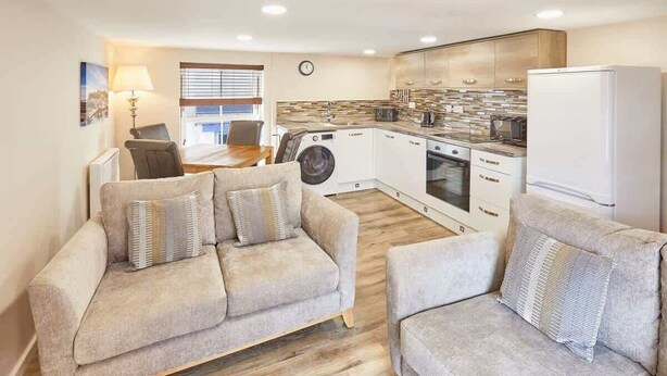 A modern apartment with a living room featuring two sofas and a dining area with a table and chairs. A kitchen with a washing machine and a refrigerator is visible. - Host & Stay | Lobster Pot Apartment (Whitby)
