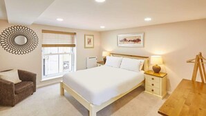 A bedroom with a large bed, bedside tables, a chair, a wooden desk, a window with blinds, and a wall decoration. - Host & Stay | Lobster Pot Apartment (Whitby)