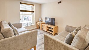 A living room with two beige sofas, a wooden TV stand, and a window with blinds. - Host & Stay | Lobster Pot Apartment (Whitby)