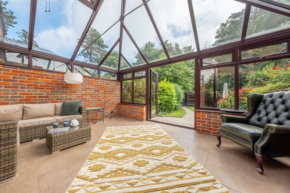 A conservatory with a brick wall, wicker furniture, a leather armchair, and a patterned rug.