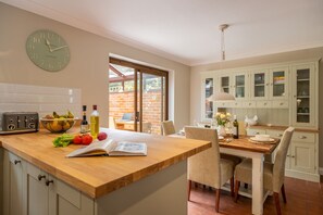 A kitchen with a wooden countertop, a clock, and a dining area with chairs and a table. - The Innings, Burnham Deepdale, Norfolk (Burnham Deepdale)