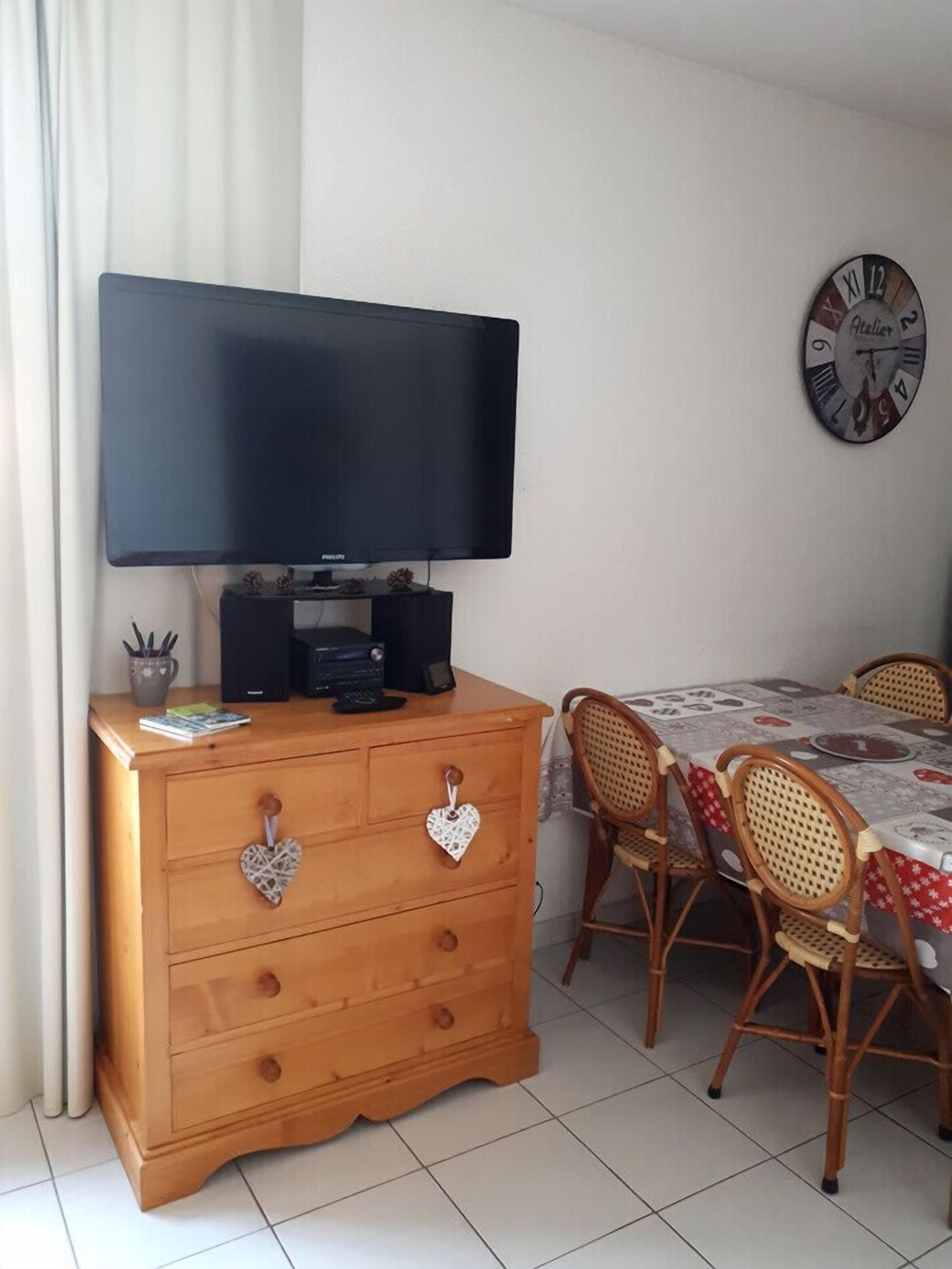 A room with a wooden dresser, a flat-screen TV, a dining table with chairs, and a wall clock.