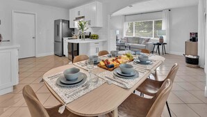 A modern kitchen with white cabinets, a stainless steel refrigerator, and a dining area with a wooden table set for four. - Steps from Beach / Quaint Cottage / Hot Tub / Surf Key / H0MES (Fort Lauderdale)