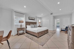 Une chambre à coucher avec un grand lit, une table de chevet, une chaise, une commode et une fenêtre avec des stores.