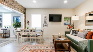Una sala de estar con un sofá verde, una mesa de centro, un comedor con mesa y sillas, un televisor montado en la pared y una pared de ladrillo decorativa.