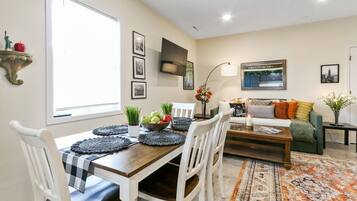 A dining area with a wooden table, white chairs, and a black and white checkered tablecloth. There is a bowl of fruit on the table and a decorative wall-mounted shelf with a statue.