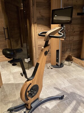 Ein stationäres Fahrrad mit Holzrahmen, schwarzem Sattel und einem digitalen Bildschirm, der an einer Wand angebracht ist.
